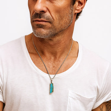 A male model wearing a white v-neck t-shirt showcasing a vibrant turquoise pendant in a chunky sterling silver setting on a box chain.
