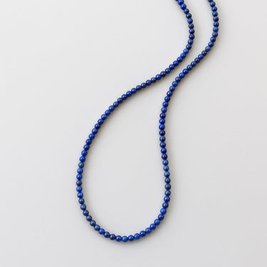A delicate strand of blue lapis lazuli gemstone beads arranged in a vertical wavy shape on a neutral background.
