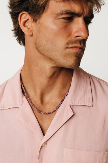 A side profile of a man wearing a peach shirt, highlighting the drape of a purple Amethyst gemstone necklace.
