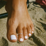 Close-up of a foot with a small silver toe ring on the second toe of the foot with white nail polish on a sandy surface