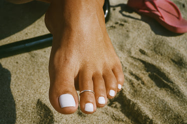 Close-up of a foot with a small silver toe ring on the second toe of the foot with white nail polish on a sandy surface