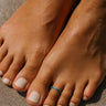 Turquoise anodized aluminum toe ring on small toe — standing on warm stone surface, COLORS Collection by Clay & Chloe Jewelry.