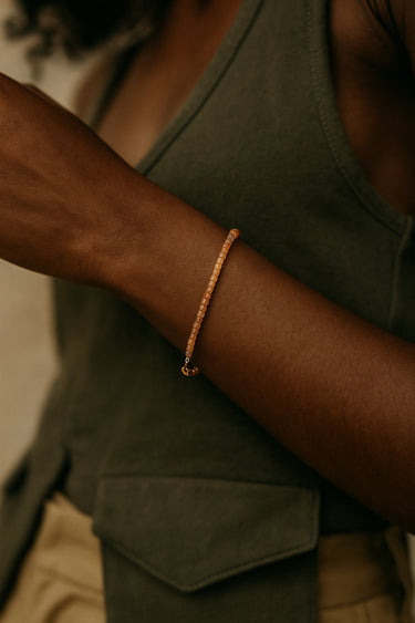A lifestyle shot of a model wearing a dainty 2mm golden sunstone cube bracelet, showcasing its warm, sparkling glow and minimalist silhouette against a casual green vest.
