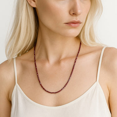 A lifestyle shot of a woman wearing the 3mm faceted garnet cube necklace, showcasing its delicate scale and clean architectural drape against a white top.
