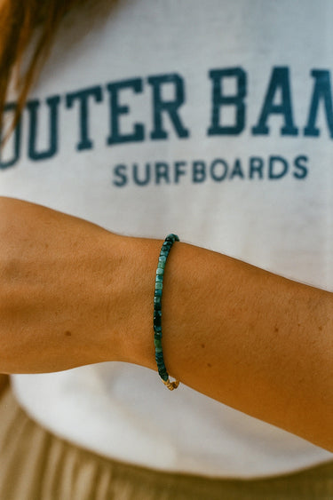 A lifestyle shot of a model wearing a dainty 3mm faceted tourmaline cube bracelet, showcasing its sophisticated sparkle and minimalist silhouette against a casual white t-shirt.
