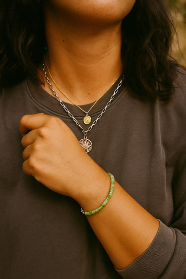 A lifestyle image of a model wearing the 4mm green chrysoprase heishi bracelet layered with silver necklaces, demonstrating its versatility as a modern staple piece with a sophisticated organic presence.
