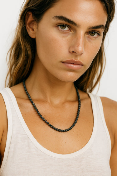 A lifestyle shot of a woman wearing the 4mm black lava necklace against a white tank top, showcasing the matte black stones and clean architectural drape.
