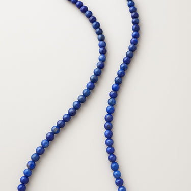 A close-up of 4mm lapis lazuli beads arranged in a fluid S-curve, emphasizing the natural matte-to-gloss texture and deep pigment of the stones.
