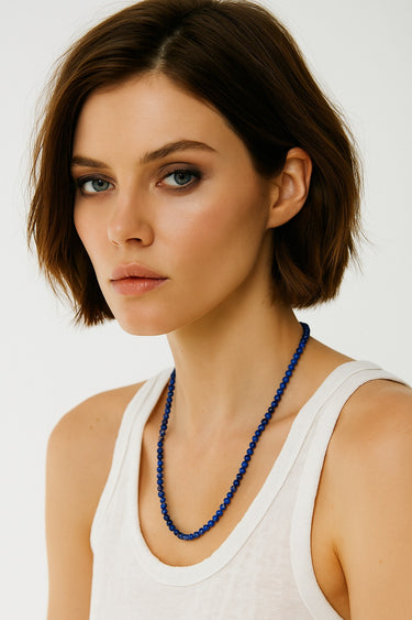 A woman wearing a 4mm lapis lazuli necklace, highlighting the vibrant blue stones and their architectural presence against a white tank top.
