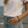 A lifestyle shot of a model wearing a 6mm black lava bead bracelet at the beach, showcasing the organic texture of the stones against a white tee and denim for a modern luxury aesthetic.
