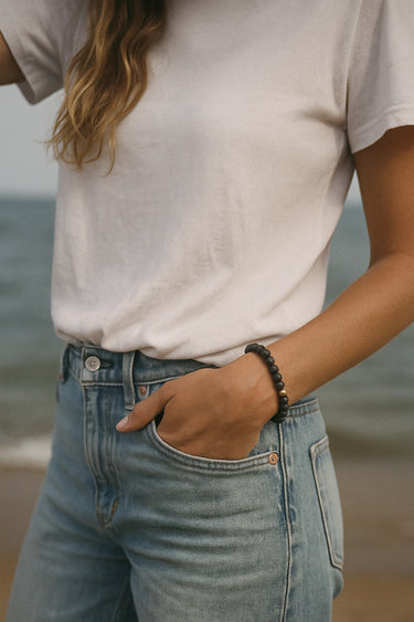 A lifestyle shot of a model wearing a 6mm black lava bead bracelet at the beach, showcasing the organic texture of the stones against a white tee and denim for a modern luxury aesthetic.
