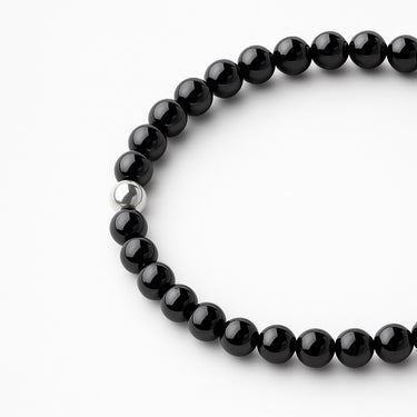 A detailed studio shot of the women's 6mm gloss black onyx bead bracelet, focusing on the seamless elastic construction and the minimalist luxury of the silver accent bead.

