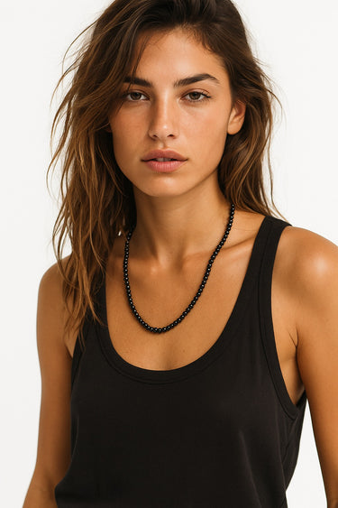 A lifestyle shot of a woman wearing the 6mm gloss onyx necklace against a black tank top, showcasing the mirror-like polish of the black stones and a clean architectural drape.
