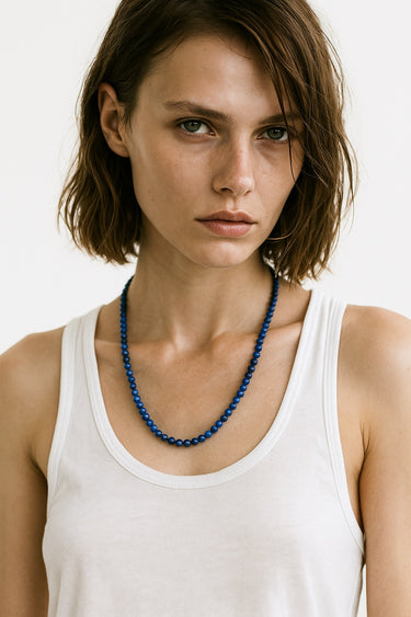 A lifestyle shot of a female model wearing a 6mm Lapis Lazuli round bead necklace against a white tank top, showcasing its deep blue color and elegant drape.
