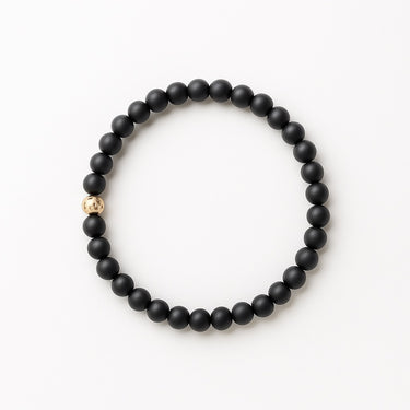 A studio hero shot of a 6mm matte black onyx bead bracelet featuring a single structural 14K gold-filled accent bead, arranged in a clean circular form on a white background.
