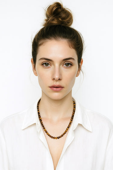 A lifestyle shot of a female model wearing a 6mm Tiger’s Eye necklace against a crisp white collared shirt, highlighting its warm tones and refined presence.
