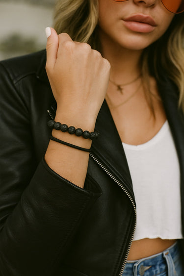 A lifestyle shot of a model wearing the 8mm black lava bead bracelet stacked with a minimalist black cuff, showcasing its architectural silhouette and modern luxury aesthetic paired with a leather jacket.
