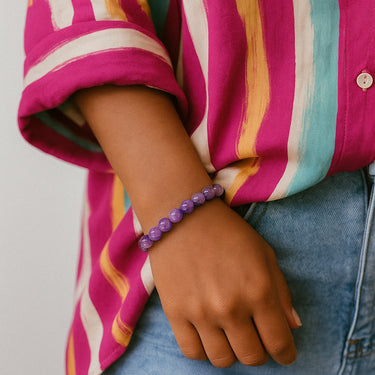 A lifestyle view of the 8mm natural amethyst bracelet on a model wearing a bright striped shirt, demonstrating how the purple gemstones add a sophisticated pop of color to a bold outfit.
