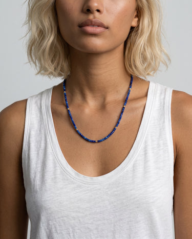 Woman wearing a white tank top and a blue lapis beaded necklace with gold accents throughout the necklace against a plain background