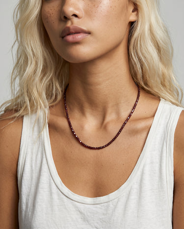 Woman wearing a white tank top and a faceted garnet beaded necklace against a neutral background. 