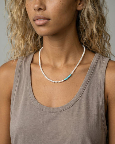 Woman wearing a handcrafted necklace featuring small white freshwater pearls and a short off-centered section of turquoise heishi disc beads, accented by a polished metal bead.