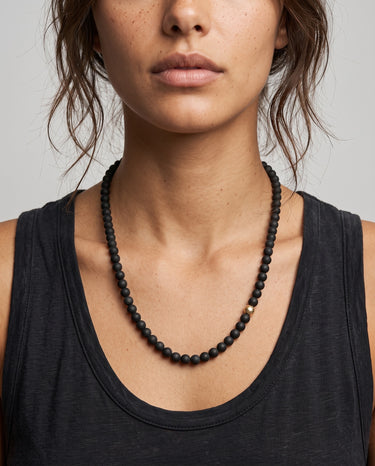 Woman wearing a black onyx beaded necklace with one single smoothe off-centered 14k gold-filled accent bead  against a neutral background.