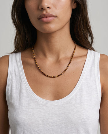 A woman wearing a white tank top and a 4mm tiger's eye beaded necklace against a neutral background