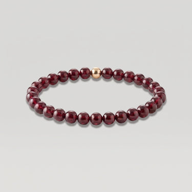 A beaded stretch bracelet featuring glossy, deep burgundy garnet round beads with a single central rose gold accent bead, displayed on a neutral light gray background.