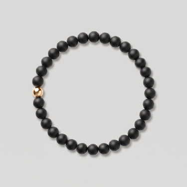 A beaded stretch bracelet made of matte black round beads with a single polished 14k gold-filled accent bead, shown on a light gray background.