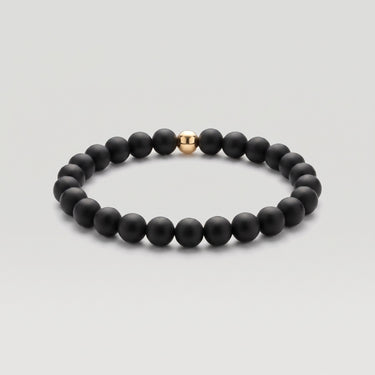 A product shot of beaded stretch bracelet made of matte black round beads with a single polished 14k gold-filled accent bead, shown on a light gray background.