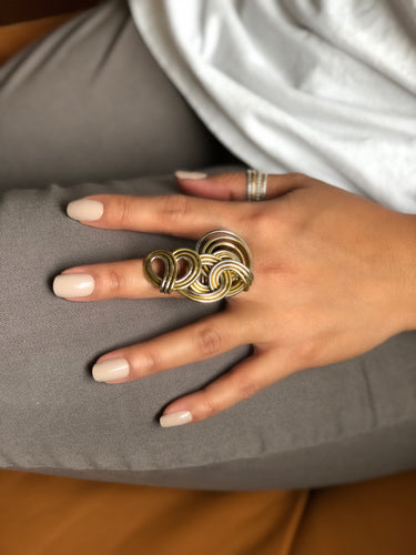 Close-up photo of a woman's hand wearing a large, statement ring on the index finger. The ring features interlocking loops of brass and sterling silver wire in an abstract, tapering spiral design.