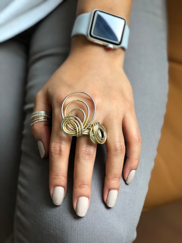 Close-up on model's hand showing the contrast between the yellow brass and white sterling silver wires, highlighting the hand-curved loops and layered construction.