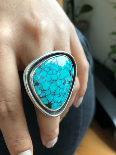 Close-up of a large sterling silver statement ring featuring a freeform turquoise stone. The stone is secured in an inner bezel, surrounded by a thick, raised outer silver wall (moat).