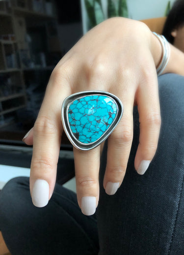 Woman wearing a large sterling silver statement ring featuring a freeform turquoise stone. The stone is secured in an inner bezel, surrounded by a thick, outer silver wall (moat).
