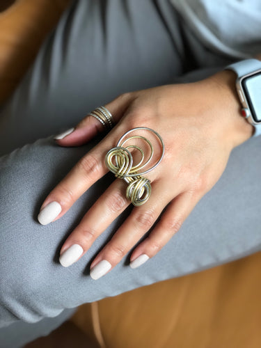 Close-up of a large mixed-metal statement ring on a person's middle finger. The design features multiple interlocking circles and spirals of brass and sterling silver wire.